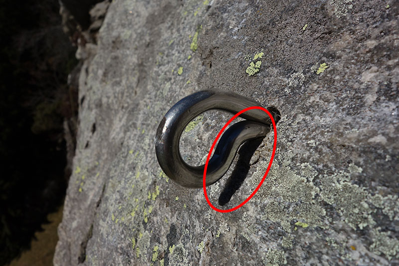 Das Bild zeigt einen schlecht angebrachten, aus dem Fels ragenden Klebehaken. Aus einer grauen Felsplatte steht der Klebehaken Schaft etwa 1 cm heraus. Die nicht gewollte Distanz der Karabineröse zum Fels ist mit einem roten Kreis markiert.
