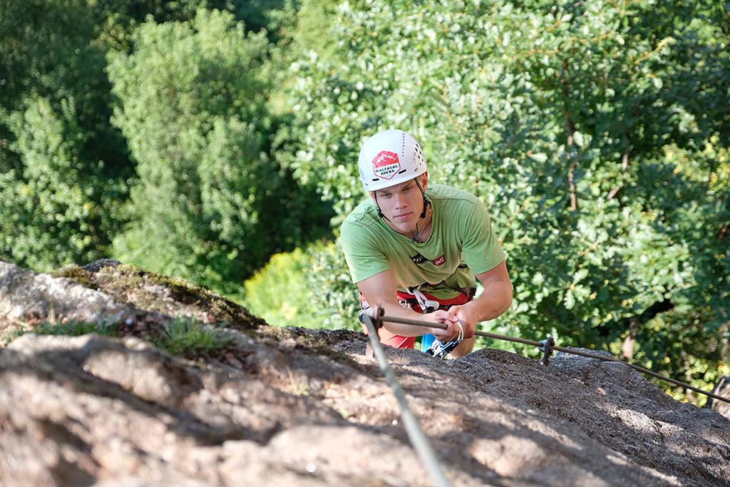 Das Bild zeigt ein erfolgreiches Beispiel für den Klettersteigbau im Tourismus, den Klettersteig Luft unter den Sohlen. Ein Mann klettert in Bildmitte ineiner steilen Passage am Stahlseil nach oben. Im Vordergrund brauner Fels, im Hintergrund grüne Blätter eines Laubwaldes