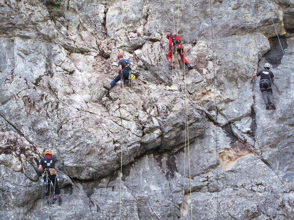 Das Bild zeigt eine graue Felswand mit vier Männern während einer Klettergarten Sanierung. Sie hängen mitten in der Wand und erledigen die Einbohr Arbeiten.