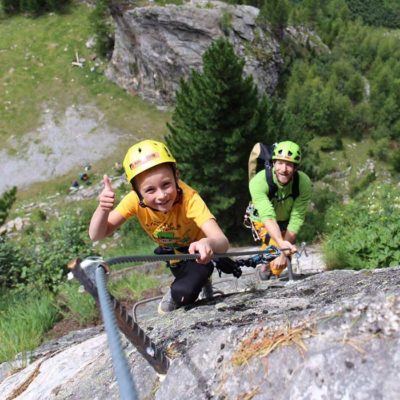 Das Bild zeigt ein Kind mit Vater am Klettersteig. Der Junge hat einen Kletterhelm Kinder Fixe auf und lächelt in die Kamera.