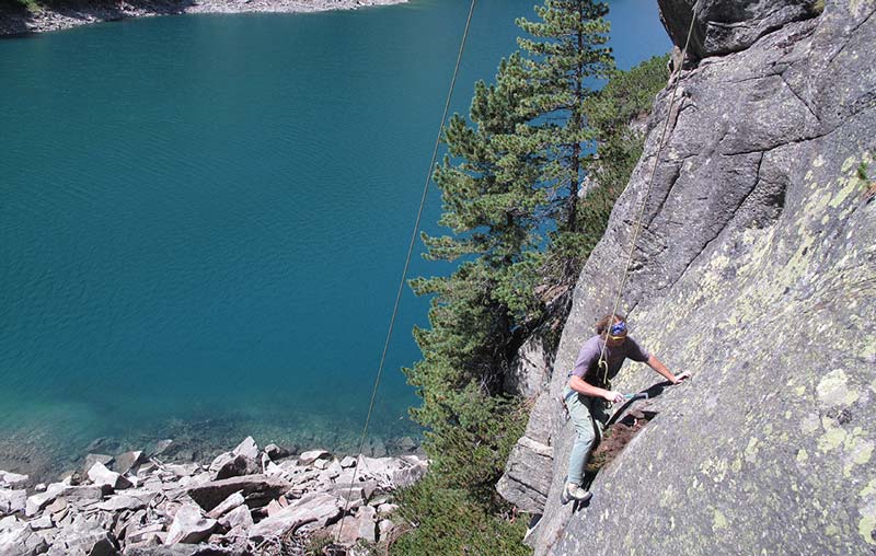 Das Bild zeigt einen Kletterer beim Putzen einer Route. Er ist in einer grauen Felswand, im Hintergrund links ein See und ein paar Bäume.