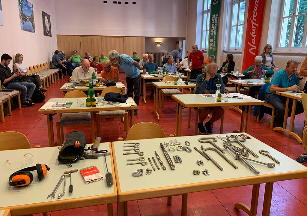 Das Bild zeigt eine Szene eines Einbohr Workshop von bolting.eu. Auf einem Tisch liegen vielerlei Einbohr Hardware, im Hintergrund dahinter viele Tische mit Workshop Teilnehmern.