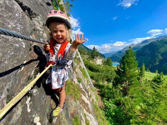 Das Bild zeigt ein dreijähriges Mädchen in einem Kleid auf einem Klettersteig. Es lacht in die Kamera und zeigt mit dem Finger nach oben. Im Hintergrund Wald, Wiese und ein Stausee sowie blauer Himmel.
