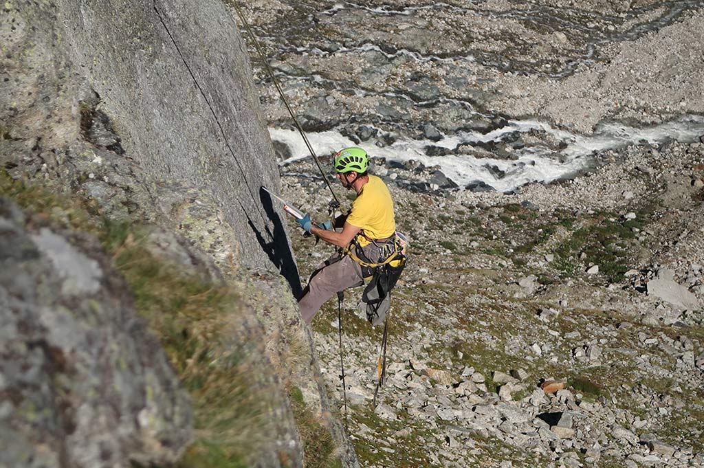 Das Bild zeigt einen Kletterer bei der Klettergarten Erschließung. Er hängt in einer grauen Felswand und klebt einen Klebehaken in die Wand. Im Hintergrund Felslandschaft und ein Gebirgsbach.