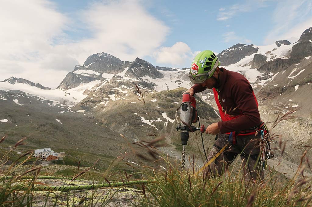 Das Bild zeigt einen Arbeiter beim Klettersteig Bau. Er steht auf einem Felsen und hält einen großen, roten Bohrhammer in den Händen. Im Hintergrund eine Hütte und eine imposante Gebirgslandschaft.