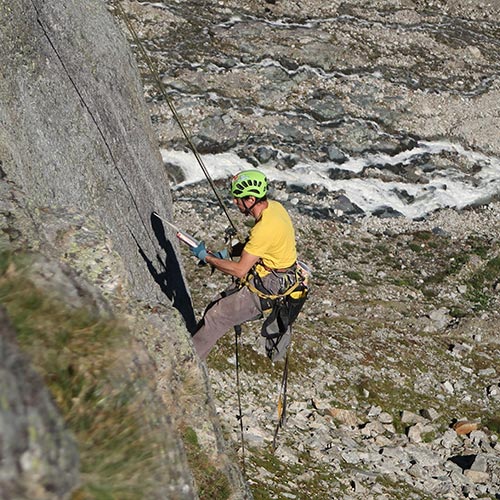 Das Bild zeigt einen Kletterer beim Einkleben eines Klebehakens in einer Kletterroute. In einer grauen Felswand hängt er am Seil und hantiert mit der Auspressppistole und dem Klebehaken. Er trägt ein gelbes T-Shirt. Im Hintergrund ein Gebirgsbach.