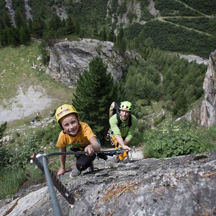 Das Bild zeigt einen Jungen der mit seinem VAter am Klettersteig unterwegs ist. Er lächelt in die Camera und hat sichtlich Spass am Sport. Im Hintergrund eine grüne Wiese, Felsen und Wald.