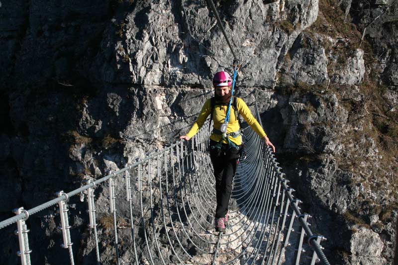 Das Bild zeigt eine Seilbrücke als Beispiel für modernen Klettersteigbau in den Dolomiten. Eine Frau balanciert über eine Drahtseilbrücke mit vielen silbernen Drahsteilen.