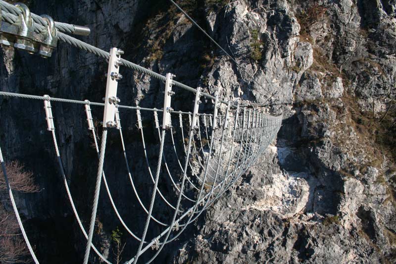 Das Bild zeigt eine Tibetan Bridge als Beispiel für modernen Klettersteigbau. Man sieht eine Drahtseilbrücke aus silbernen Seilstücken über eine Schlucht.