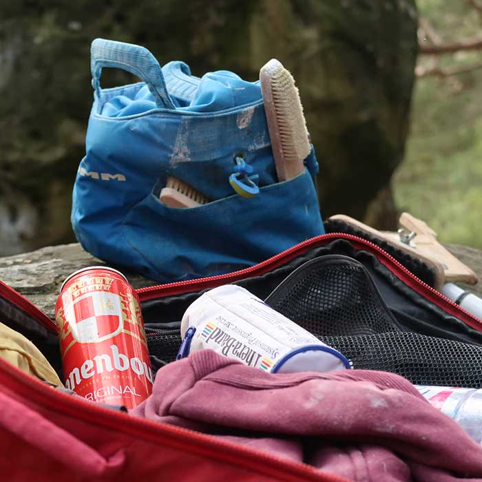 Zu sehen ist ein Bild zum Thema Chalkbag Bouldern. Im Vordergrund sieht man einen geöffneten Kletterrucksack mit einer Dose Bier und einem Theraband, im Hindergrund ein Boulder Chalkbag.