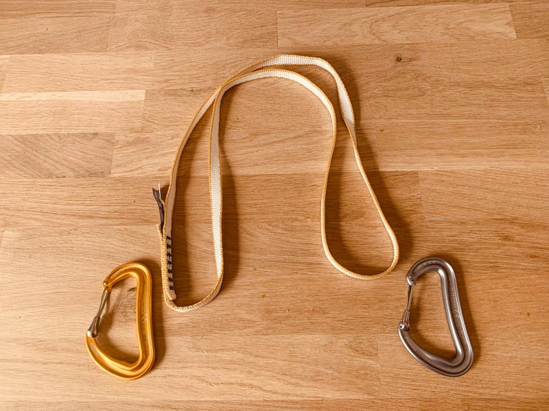 Das Bild zeigt die MAterialien für den BAu einer verlängerbaren Expressschlinge - verlängerbaren Alpinschlinge. Eine Bandschlinge und zwei KArabiner auf einer Holzplatte.