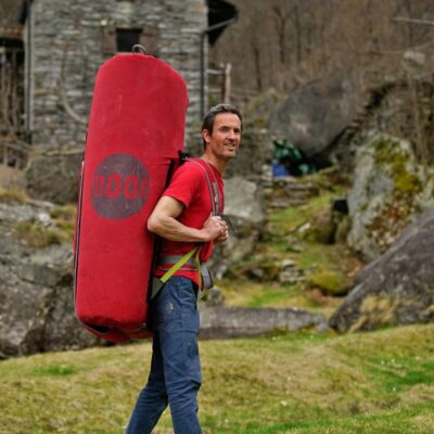 Das Bild zeigt Ben Moon mit einer roten Bouldermatte am Rücken auf einer Wiese. Im Hintergrund ein Steinhaus.