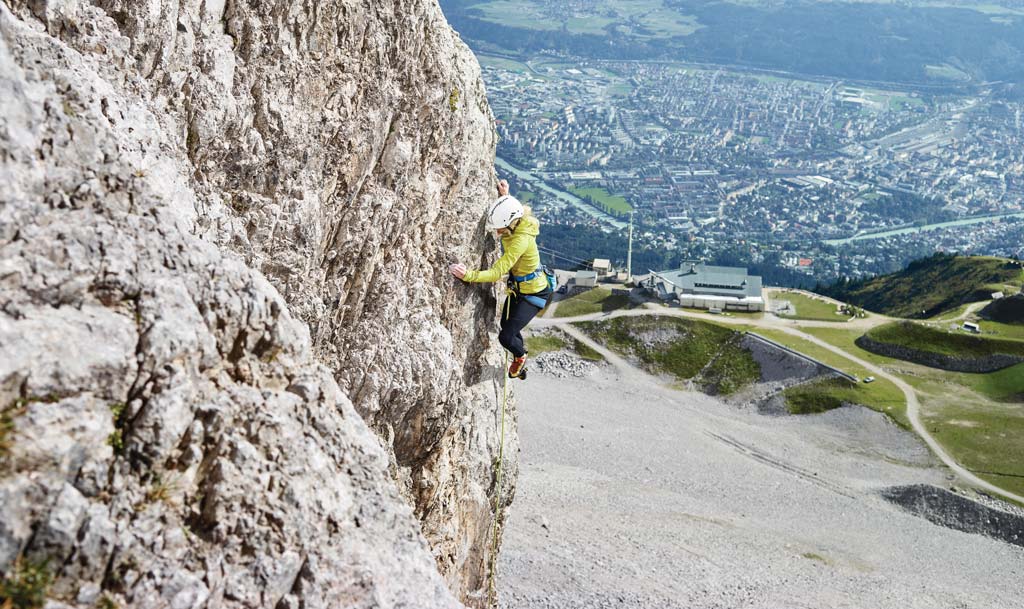 Klettererin hoch über der Stadt Innsbruck an einer grauen Felswand. Tief unten die Stadt.