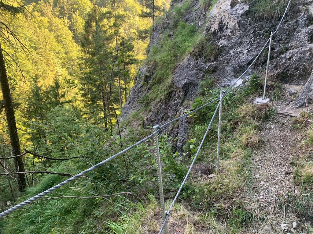 Das Bild zeigt eine Absturzsicherung auf einem Wanderweg als BEispiel für alpinen Wegebau. Silberne GEländestangen und Drahtseil sind vor einem Abgrund.