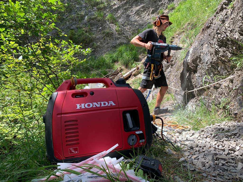Das Bild zeigt einen Arbeiter beim alpinen Wegebau, er arbeitet mit einem Schlagbohrer hinter einem roten Stromaggregat.
