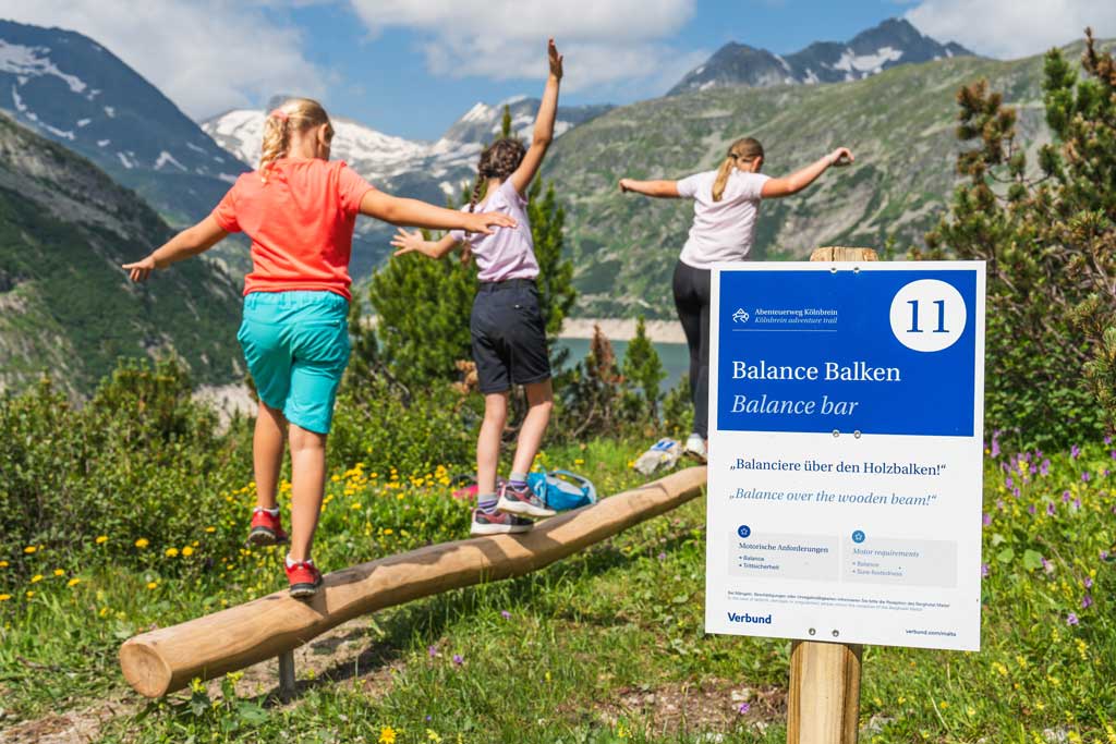 Drei Kinder auf einem Balance Balken am Abenteuerweg Kölnbrein. Im Hintergrund alpine Landschaft.