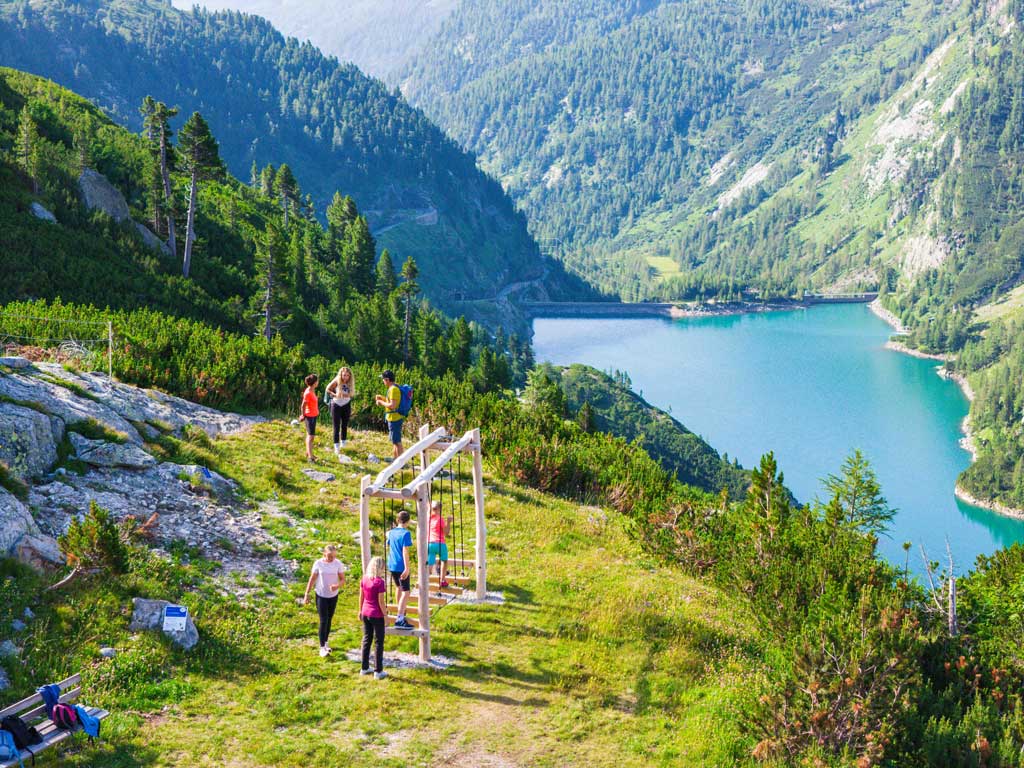 Kinder auf einer der Stationen des Abenteuerweg Kölnbrein in alpiner Landschaft und mit Stausee im Hintergrund.