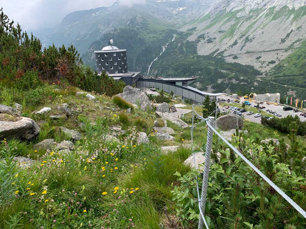 Wegebau in alpinem Gelände mit Geländerversicherung. Im Hintergrund ein Hotel und Berglandschaft.