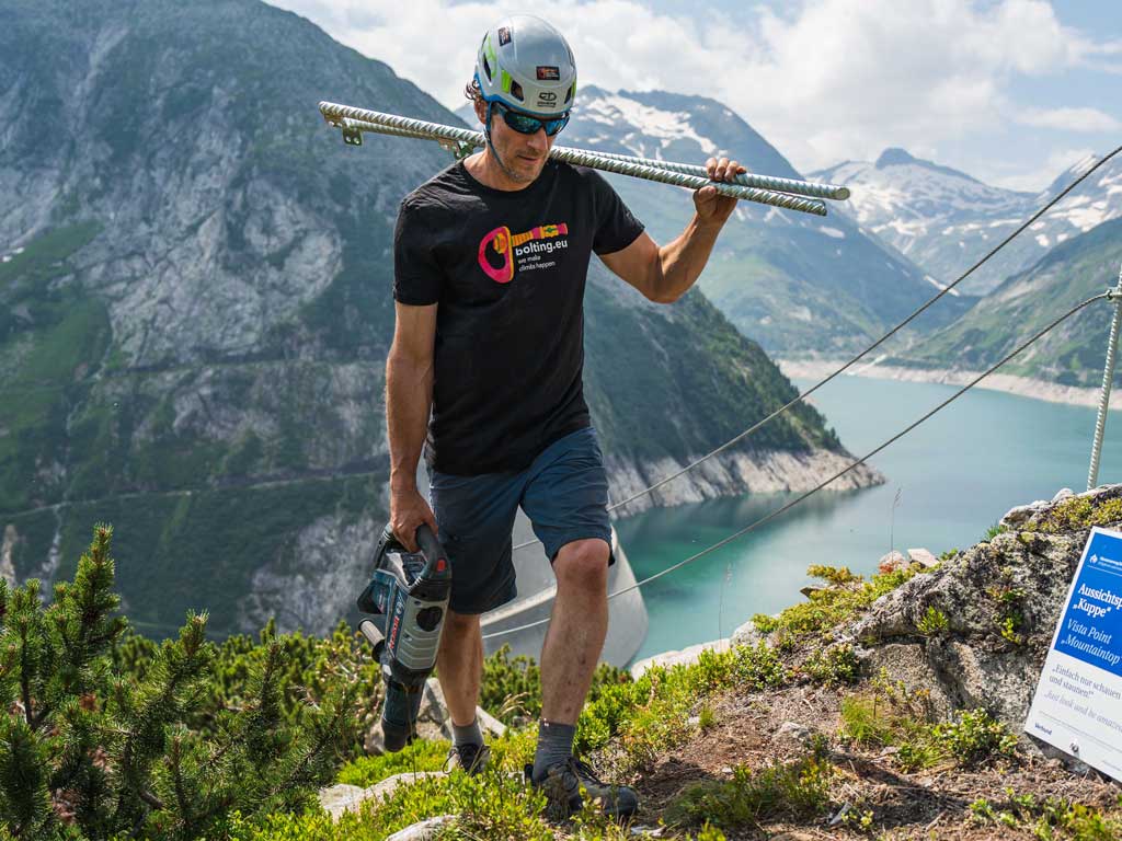 Ein Mann beim Tragen von Ausrüstung für einen Klettersteigbau in alpiner Landschaft mit Stausee.