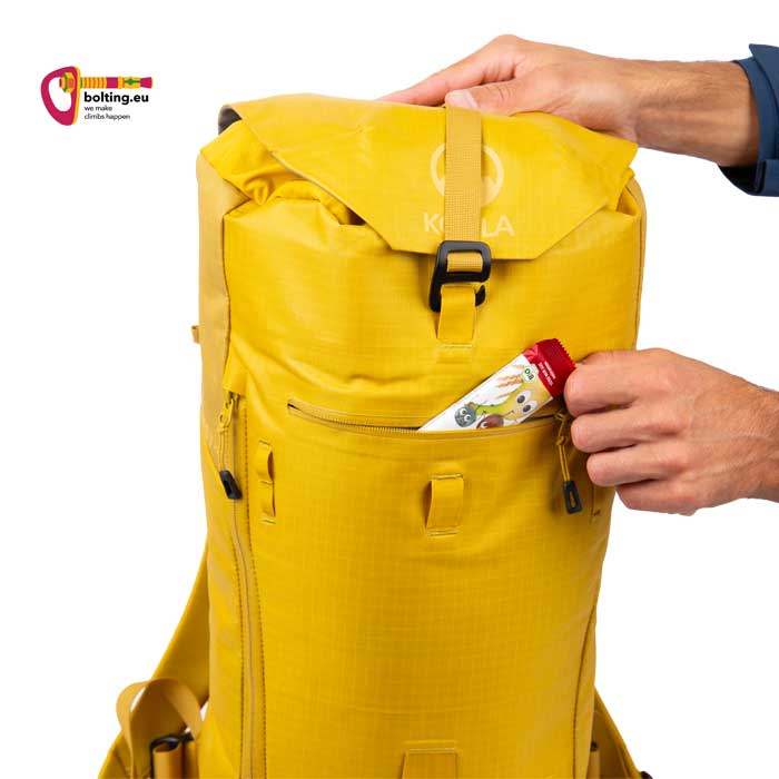 Hand steckt einen Müsliriegel in die Außentasche eines gelben Kletterrucksacks.